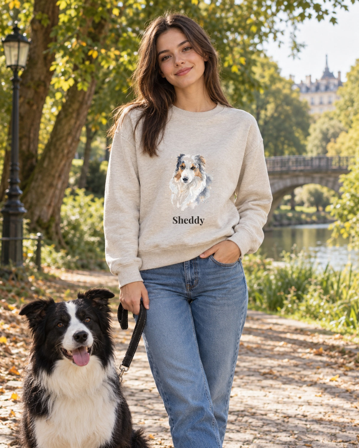 Frau im hellen Sweatshirt mit Hundeportrait im Aquarell-Stil und Namen Sheddy, im Park mit Border Collie