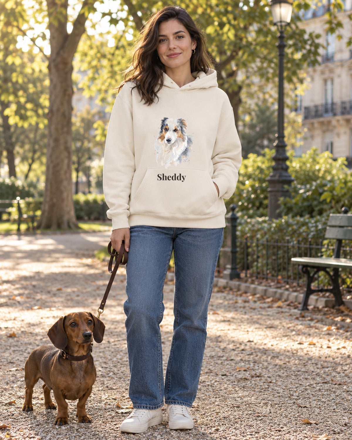 Frau im cremefarbenen Hoodie mit Hundeportrait im Aquarell-Stil und Namen Sheddy, im Park mit Dackel an der Leine