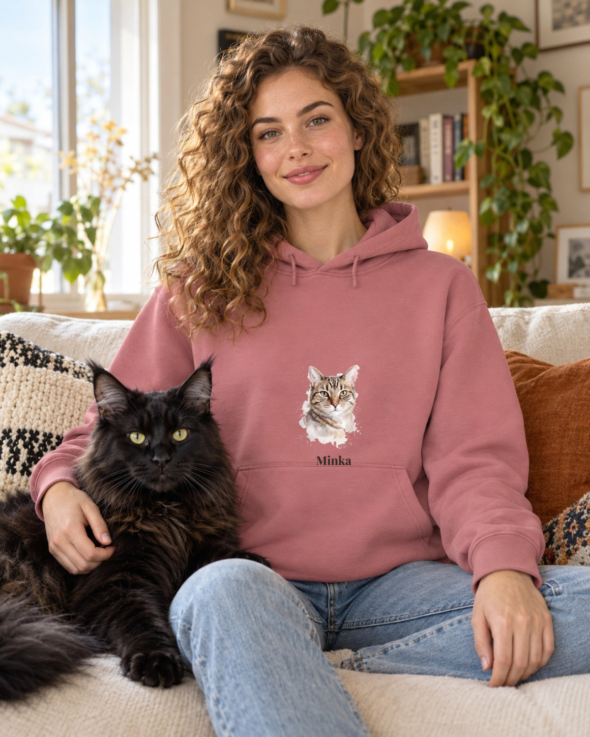 Frau im rosa Hoodie mit Katzenportrait im Aquarell-Stil und Namen Minka, auf dem Sofa neben einer schwarzen Maine-Coon-Katze