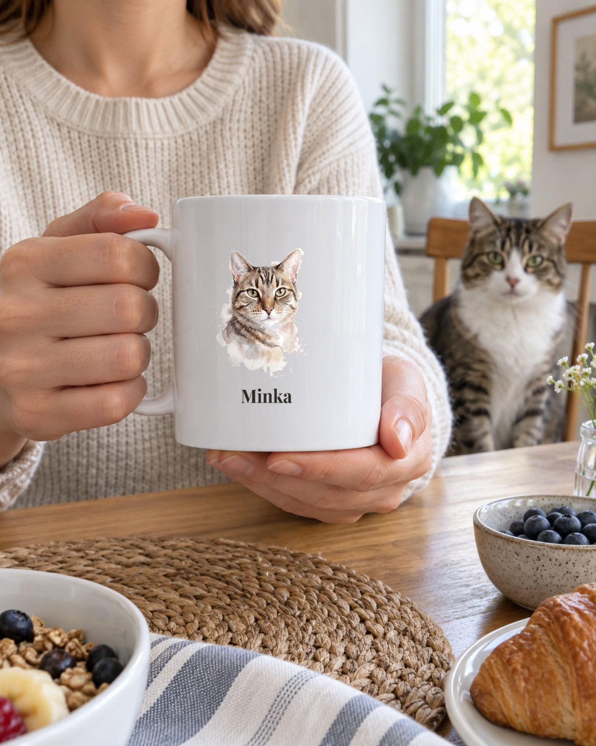 Weiße Tasse mit Katzenportrait im Stil Aquarell und dem Namen Minka am Frühstückstisch, eine Katze sitzt auf dem Stuhl dahinter