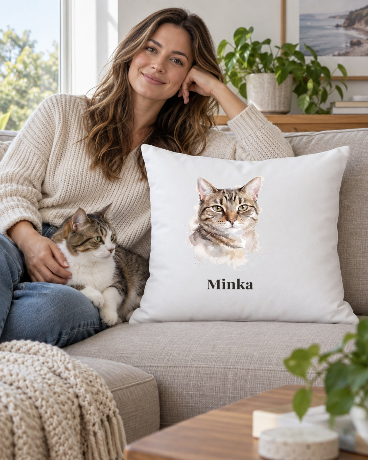 Weißes Kissen mit Katzenportrait im Stil Aquarell und dem Namen Minka auf dem Sofa, eine Frau mit ihrer Katze daneben