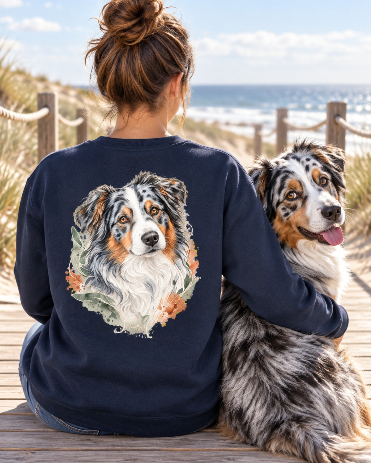 Frau im navyblauen Sweatshirt mit Australian Shepherd im Aquarell-Stil auf dem Rücken, daneben ihr Hund am Strand