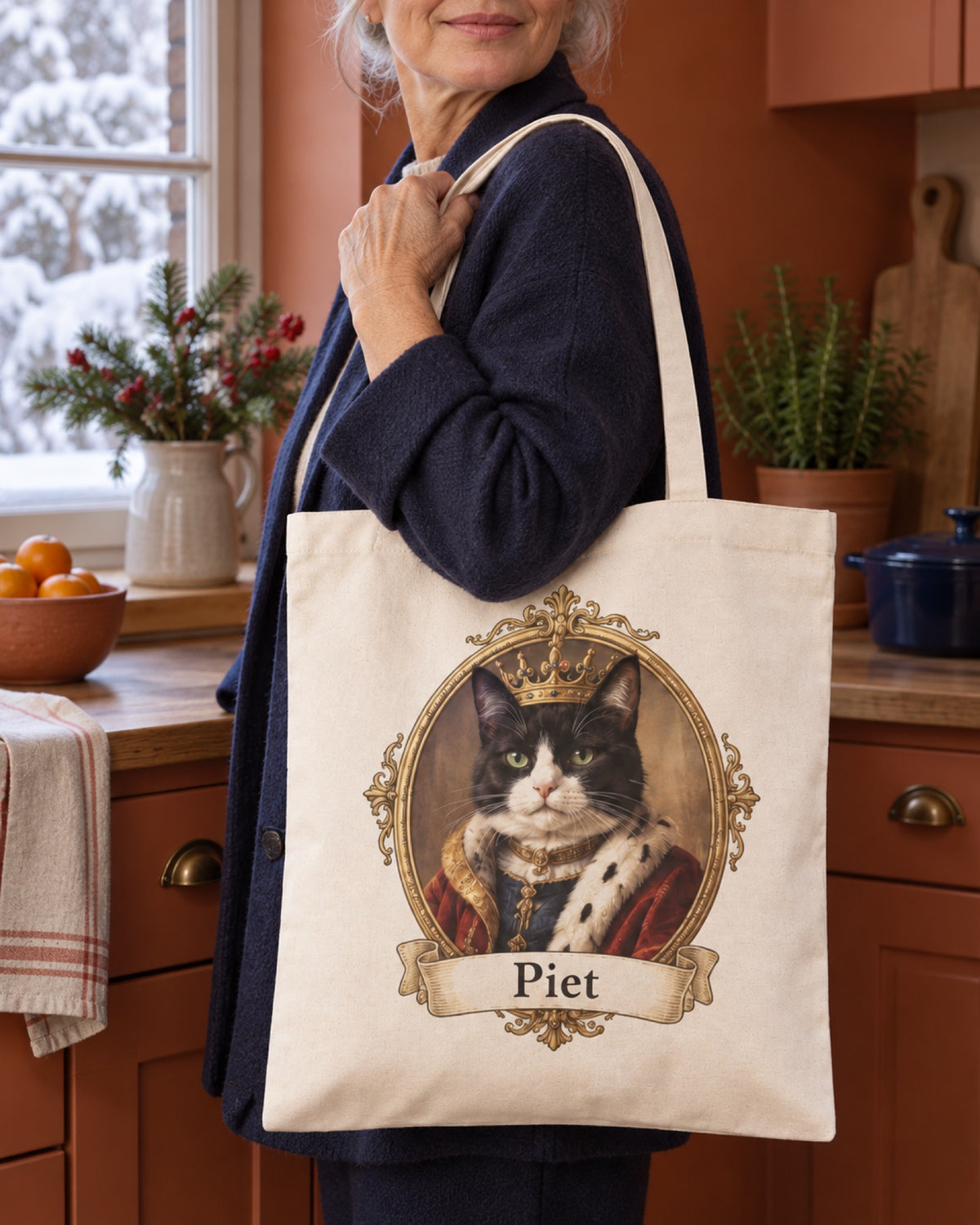 Naturfarbene Baumwolltasche mit schwarzweißer Katze im Royal Stil und Namen Piet auf der Hand einer älteren Frau mit dunkelblauer Jacke in einer terrakottafarbenen Küche am Winterfenster