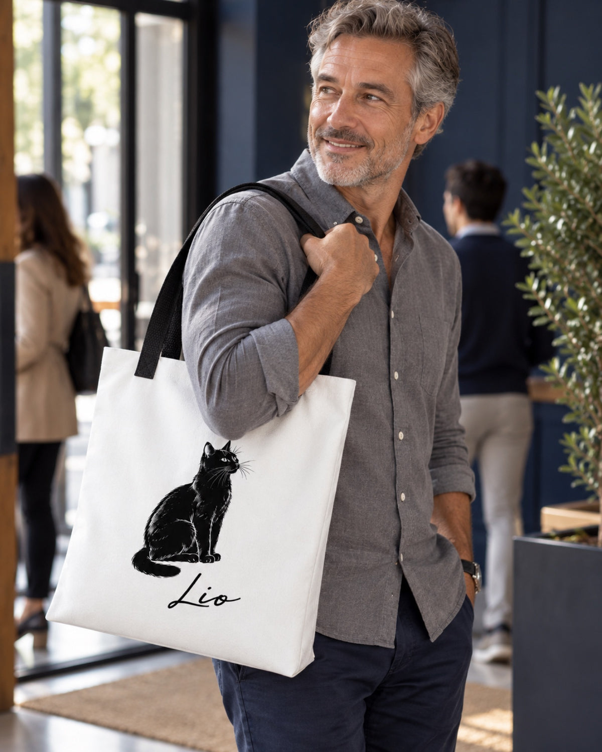 Weiße Stofftasche 39 × 39 cm mit schwarzen Henkeln, schwarze Katze Portrait und Namen Lio an der Schulter eines älteren Mannes mit grauem Hemd in einem dunkelblauen Eingangsloft